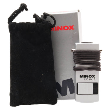 Minox MD 6x16 monocular with grey lanyard wrapped around body, black velvet pouch, and original Minox box